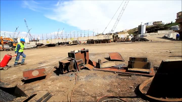 A worker in hi-vis wheels a gas cylinder across a ship dismantling yard, with cranes behind.